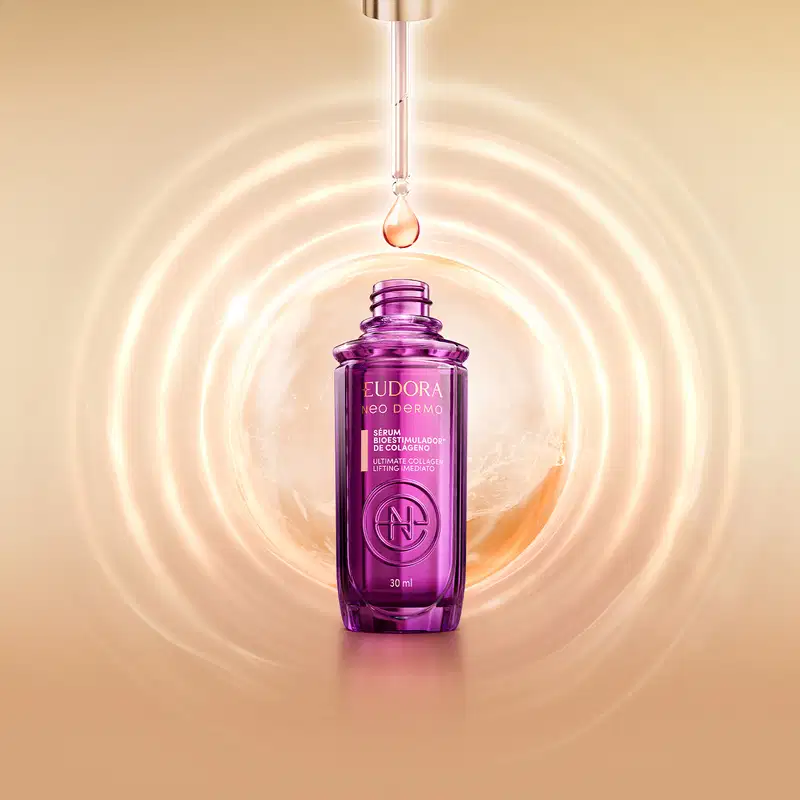 Sérum Bioestimulador de Colágeno. O frasco possui um formato cilíndrico e é predominantemente na cor roxa, com detalhes em dourado. O frasco está suspenso em um fundo desfocado, com o conta-gotas aparecendo e deixando cair uma gota.