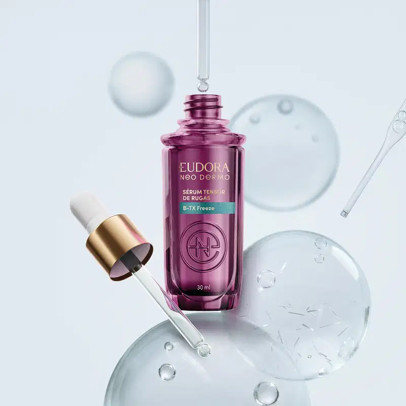 Frasco roxo com rótulo da marca Eudora Neo Dermo, com a tampa em conta gotas ao lado. Ao redor, temos espécie de gotas lisas, translúcidas e brilhantes, indicando a textura do produto.