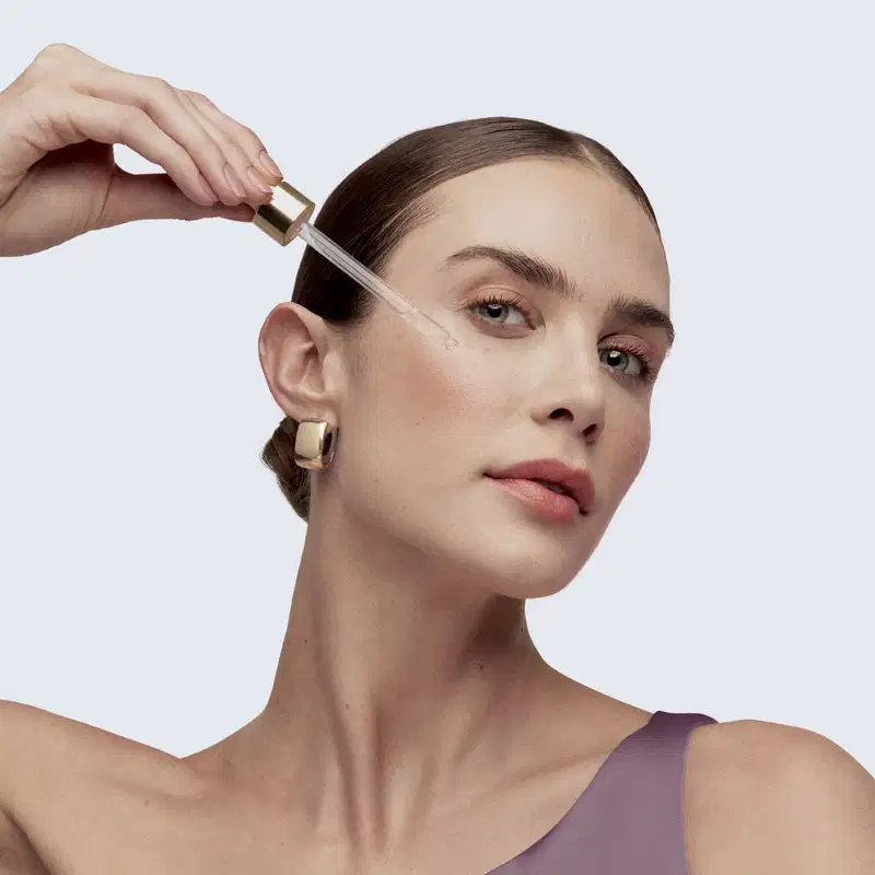 Uma mulher aplica um produto de skincare em seu rosto, com uma conta gotas transparente, com tampa dourada em primeiro plano.