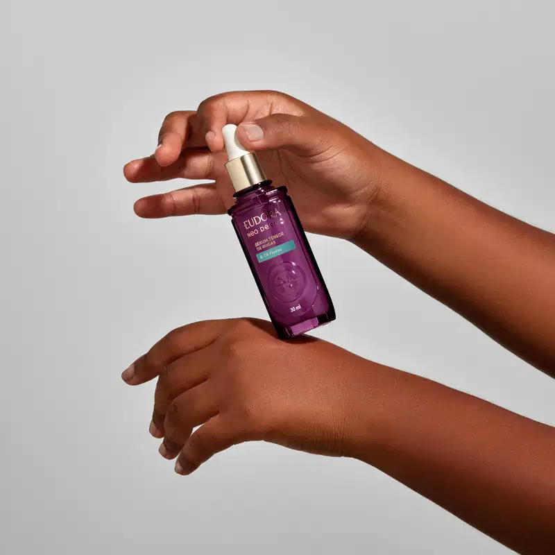 Frasco roxo com rótulo dourado da marca Eudora Neo Dermo, com uma tampa em conta gotas. O produto está sendo segurado por mãos femininas.