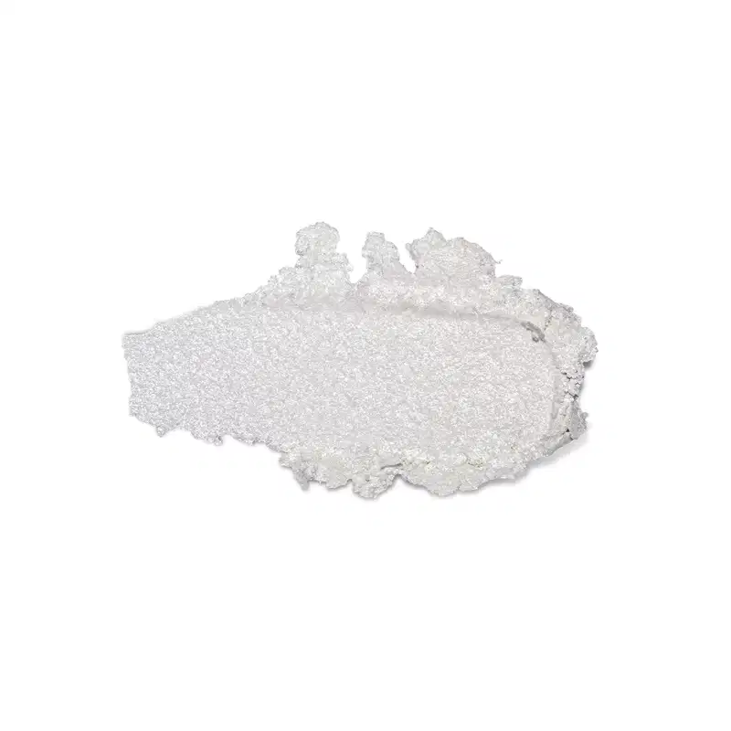 Textura do produto Iluminador Glam. Trata-se de um aglomerado de cristais brancos e brilhantes, com uma textura granulada e irregular. Não há embalagem ou rótulo visível na imagem, apenas o próprio produto.