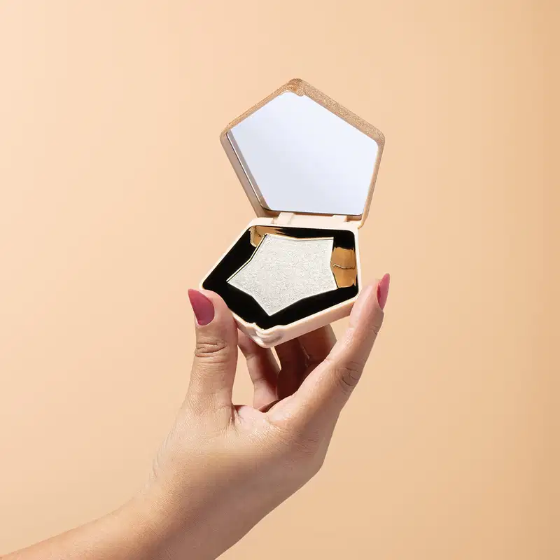 O produto apresentado é um estojo de maquiagem em formato pentagonal, com tampa espelhada. A embalagem é de cor clara, com detalhes dourados, dando um aspecto elegante e sofisticado. O estojo contém um iluminador, com uma textura brilhante e cintilante.