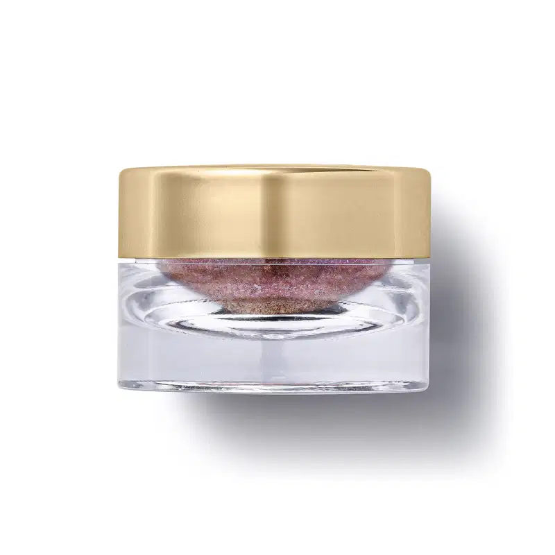 O produto apresentado é um pote transparente com uma tampa dourada. Dentro do pote, há uma substância de cor rosa. A embalagem possui um design elegante e minimalista, com a tampa dourada contrastando com o transparente.