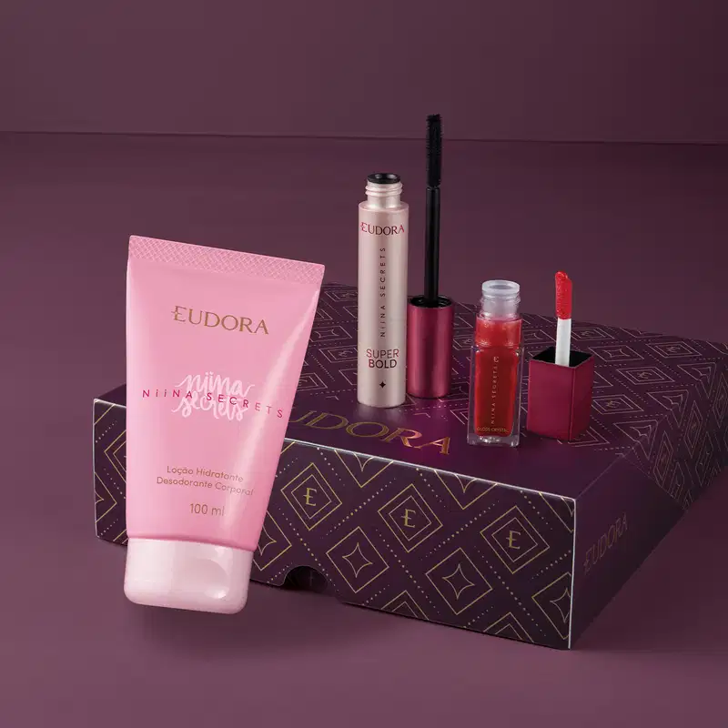A caixa roxa e dourada da marca Eudora contém diversos produtos de beleza, incluindo uma loção hidratante corporal da linha Nina Secrets, um gloss que está aberto e um frasco de máscara de cílios preta também com o aplicador ao lado.