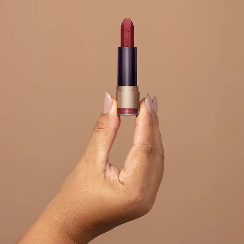 Embalagem de batom vermelho da marca Eudora Make sobre superfície bege, com mão segurando o produto em destaque.
