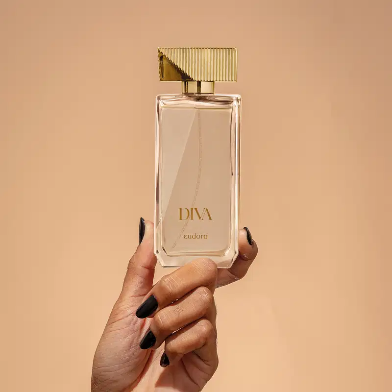 Frasco do Desodorante Colônia Diva da marca Eudora, com design elegante e sofisticado sendo segurado por uma mão feminina. Embalagem de vidro transparente com tampa dourada e detalhes verticais. 