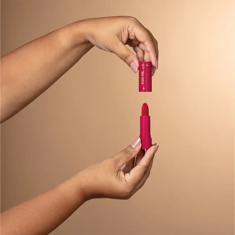 A imagem mostra um batom vermelho em formato de tubo, com a marca "Eudora" impressa no produto. Uma mão está segurando o produto aberto, coma tampa na parte de cima, mostrando o formato interno do batom.