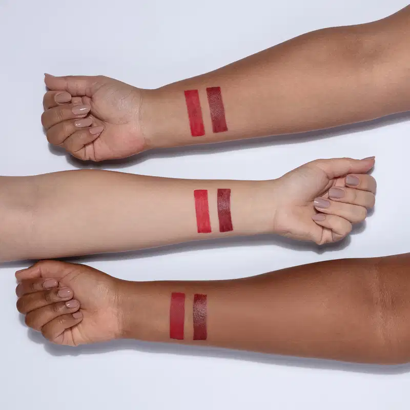 Imagem de 3 braços com peles de cores diferentes mostrando a aplicação de dois batons na cor vermelha. Cada batom possui duas barras verticais de cor vermelha, provavelmente com diferentes tonalidades ou acabamentos, aplicados no antebraço da pessoa.