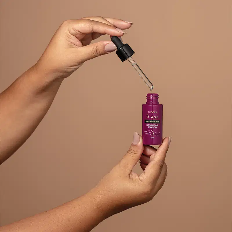 O Potencializador de Nutrição da linha Siàge Pro Cronology está sendo segurado por uma mão, aberto. O frasco tem um conta-gotas preto na tampa. Parece ter uma consistência líquida e é apresentado em um ambiente com fundo bege.