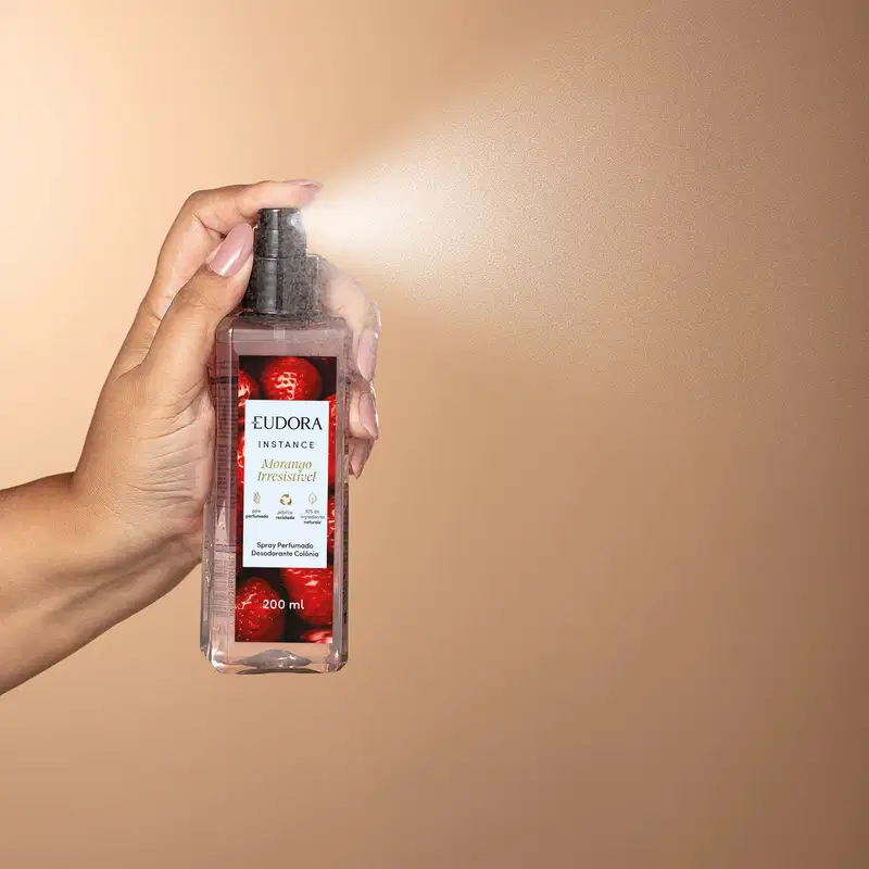 O produto mostrado é um spray perfumado da marca Eudora, que está sendo segurado por uma mão feminina. O borrifador está acionado, saindo o produto em forma de spray. O frasco é transparente com uma tampa preta e um rótulo vermelho e branco.