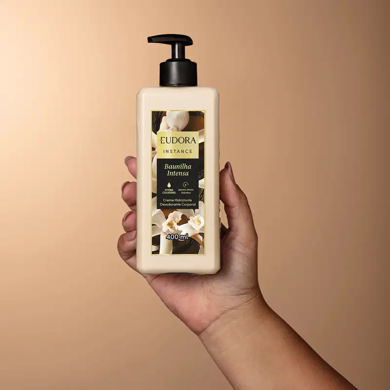 Imagem de um produto de hidratação corporal da marca Eudora, com embalagem branca e preta, em um ambiente com fundo bege, sendo segurado por uma mão.