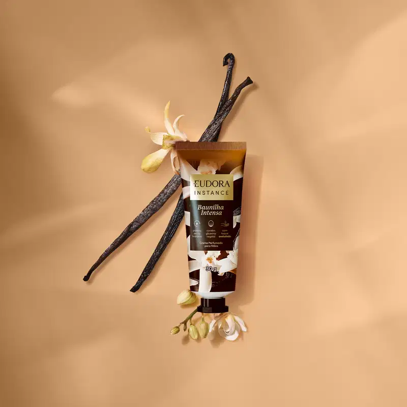 Ambiente de fundo com tons terrosos, apresentando um tubo de creme de mãos perfumado da marca Eudora Instance, com flores e vagens de baunilha ao redor, sugerindo um contexto de cuidados corporais e fragrância.