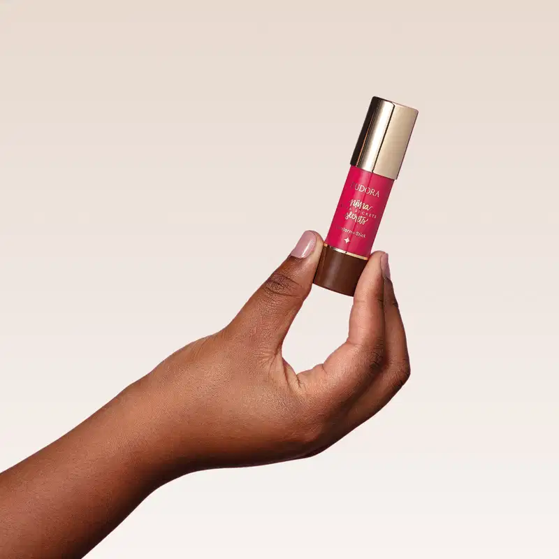 O produto mostrado na imagem é o Contorno Stick de Niina Secrets, que possui uma embalagem cilíndrica dourada com um rótulo rosa brilhante. O produto está sendo segurado por uma mão com pele escura.