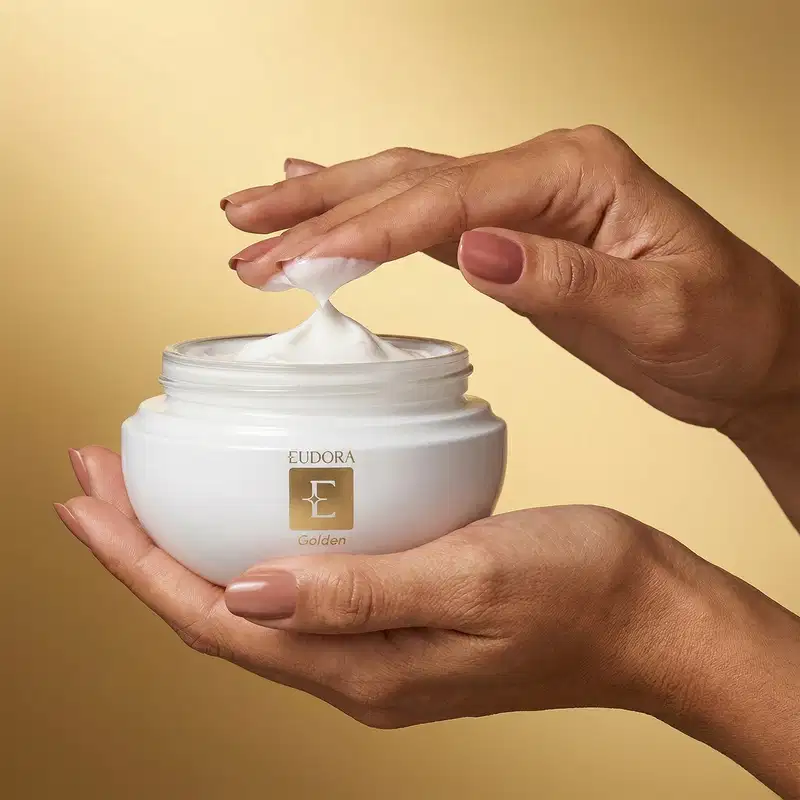 Mãos segurando um pote de creme da marca Eudora Golden em um fundo bege/dourado, sugerindo um ambiente de cuidados de beleza e cosmética.