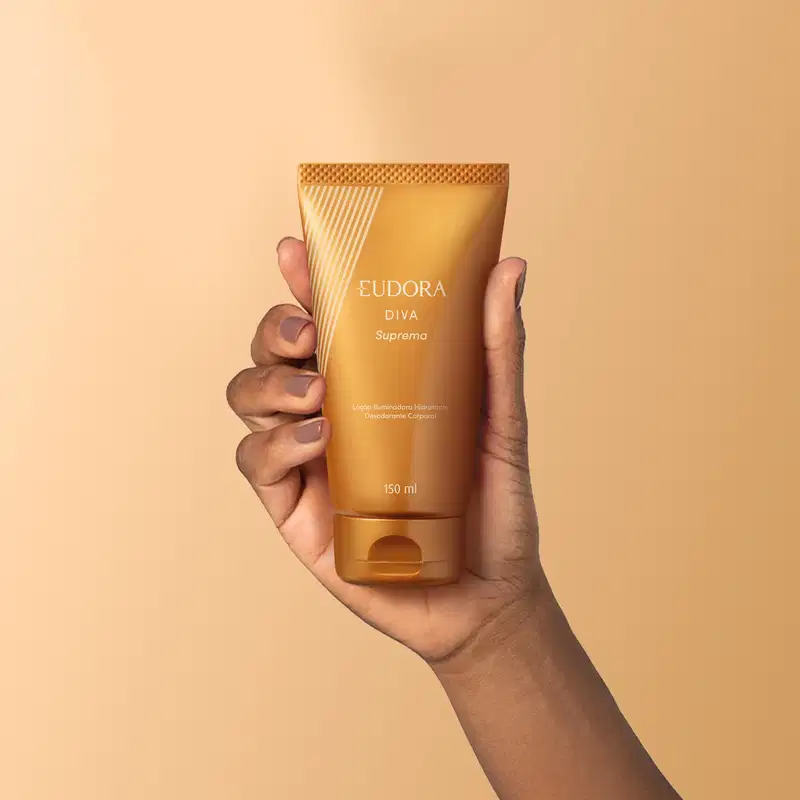 O produto apresentado é um desodorante corporal da linha Diva Suprema. O recipiente é um tubo dourado com detalhes em relevo e o nome da linha "Diva Suprema" impresso. O tubo possui 150 ml de volume e é descrito como um loção iluminadora hidratante.