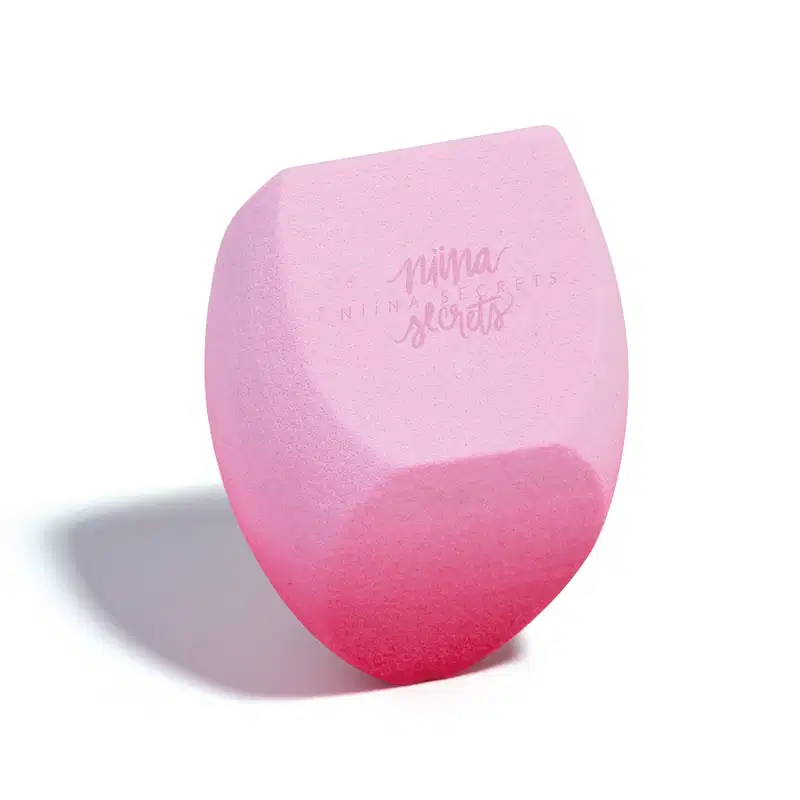 O produto apresentado é uma esponja para maquiagem da marca Niina Secrets, com uma coloração degradê de rosa claro a rosa escuro. Possui um formato arredondado e achatado em diferentes partes. Em outro ângulo.