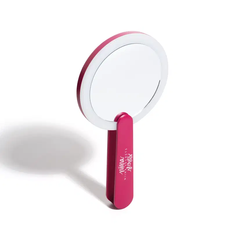 O produto apresentado é um espelho de mão da marca Niina Secrets. Possui um formato circular com um aro branco e um cabo de cor rosa vibrante. O nome da marca "Niina Secrets" está impresso no cabo do espelho em letras brancas.