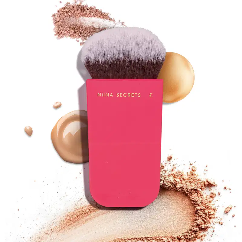 O produto apresentado é um pincel de maquiagem da marca Niina Secrets. O pincel possui cerdas macias e uma base em um tom rosa vibrante, com o nome da marca impresso em letras douradas. Texturas em pó, líquidas e cremosas estão dispostas no fundo.