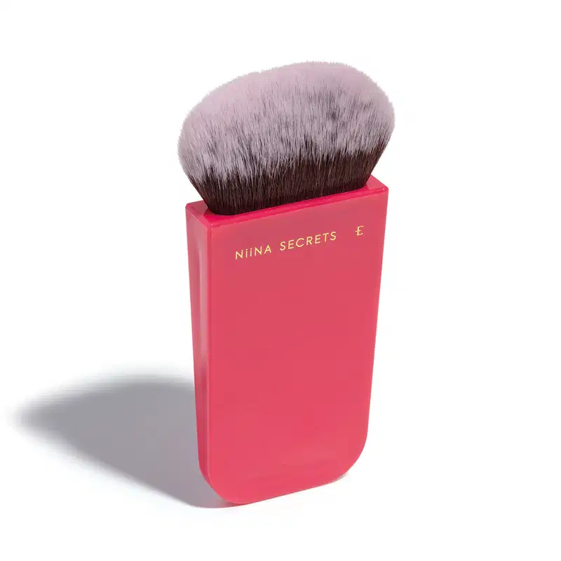 O produto apresentado é um pincel de maquiagem da marca Niina Secrets. O pincel possui cerdas macias e uma base em um tom rosa vibrante, com o nome da marca impresso em letras douradas. Em outro ângulo.