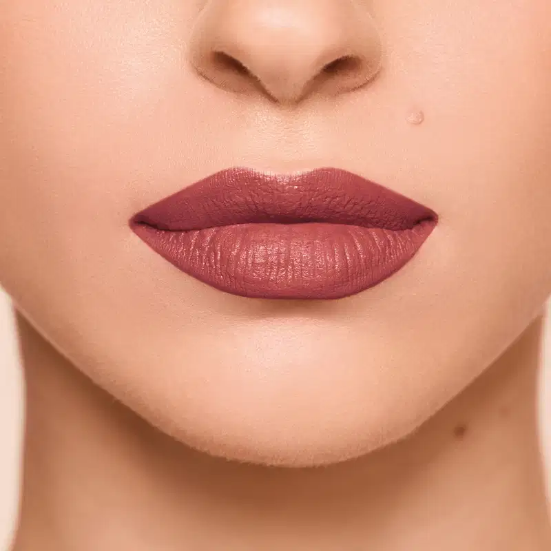 Imagem da aplicação do batom em lábios de pessoa de pele branca. O tom do batom apresenta-se em marrom claro rosado nude, elegante, sóbrio e com alta pigmentação e cobertura cremosa matte uniforme.
