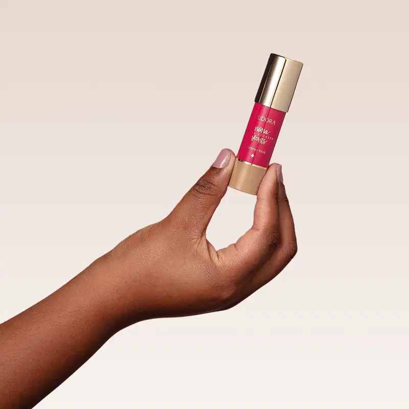 O produto mostrado na imagem é o Contorno Stick de Niina Secrets, que possui uma embalagem cilíndrica dourada com um rótulo rosa brilhante. O produto está sendo segurado por uma mão com pele escura.