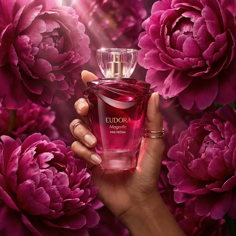 A mão segura um frasco do desodorante colônia Eudora Magnific Pink Peonia, cercado por flores de peônia rosa em um ambiente elegante, destacando a sofisticação e delicadeza do produto.