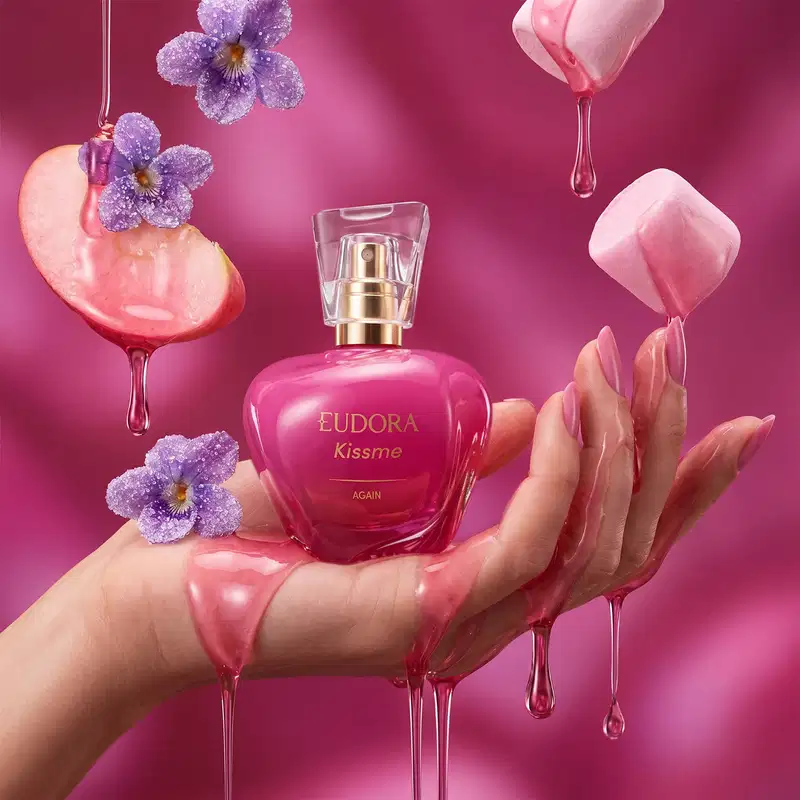 Frasco de item de perfumaria Eudora Kiss Me Again em mãos sobre fundo rosa com gotas, maçã, marshmallow e flores violetas decorativas.
