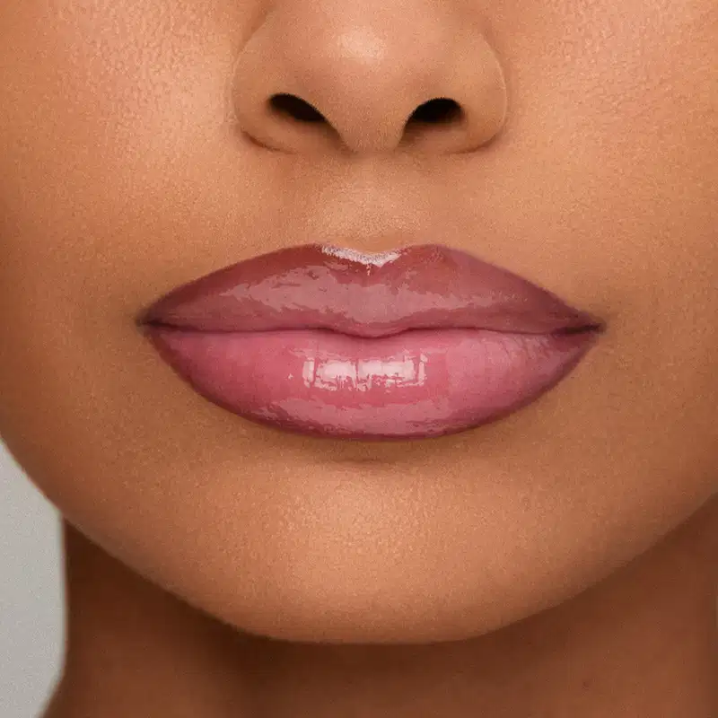 Imagem em close-up da modelo, mostrando o efeito do Gloss Labial: Lábios rosados com brilho, em primeiro plano, sobre fundo neutro.
