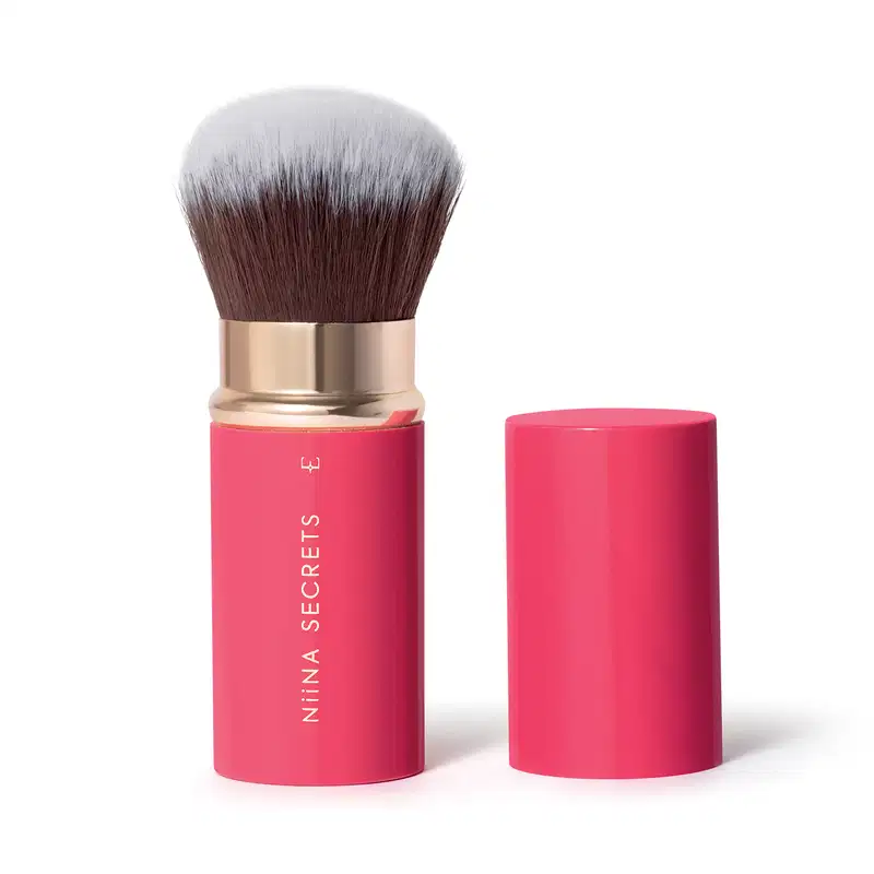 Pincel de maquiagem com cerdas marrons e cabo dourado e rosa. Recipiente cilíndrico rosa com rótulo branco e texto "Nina Secrets". O item está aberto e tem a tampa, também rosa, ao lado.