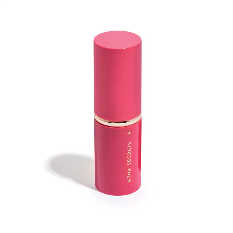 Recipiente cilíndrico rosa com tampa dourada. Rótulo com o texto "Nina Secrets" em letras douradas. Trata-se de um pincel retrátil com tampa.