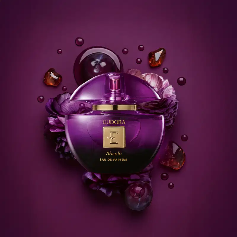 Frasco de perfume Eudora Absolu em tom roxo, com tampa dourada e rodeado por flores roxas e gotas de líquido âmbar. Embalagem elegante e sofisticada.