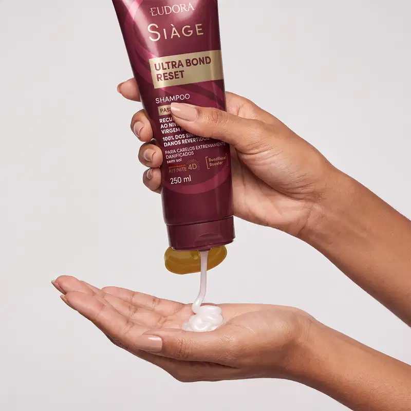 Tubo de shampoo para cabelos da marca Siàge, em tons de vinho e dourado. Uma mão está apertando a bisnaga e está saindo o produto, com textura cremosa e brilhante.