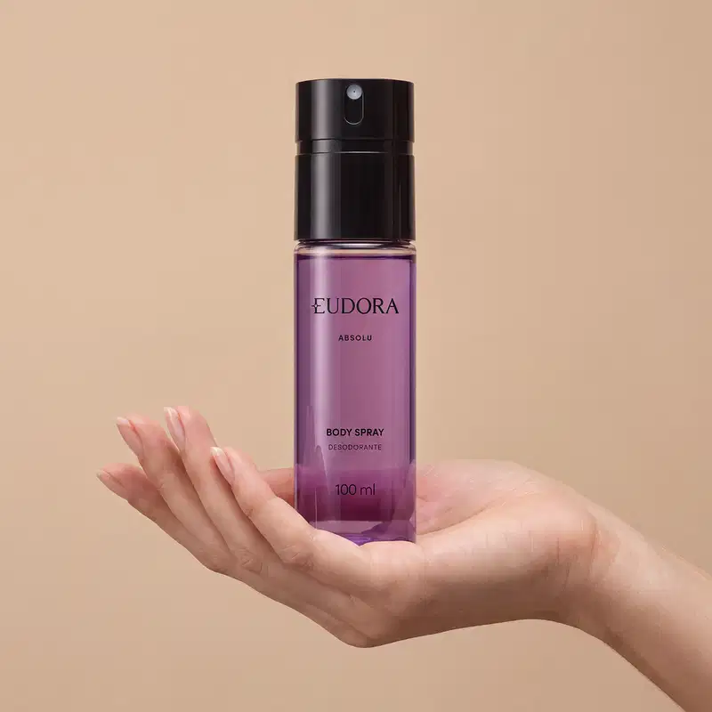 Frasco preto de item de perfumaria da marca Eudora, com líquido roxo claro, escrito: ABSOLU BODY SPRAY DESODORANTE em letras pretas. O produto está sendo segurado por mão feminina sobre fundo bege.