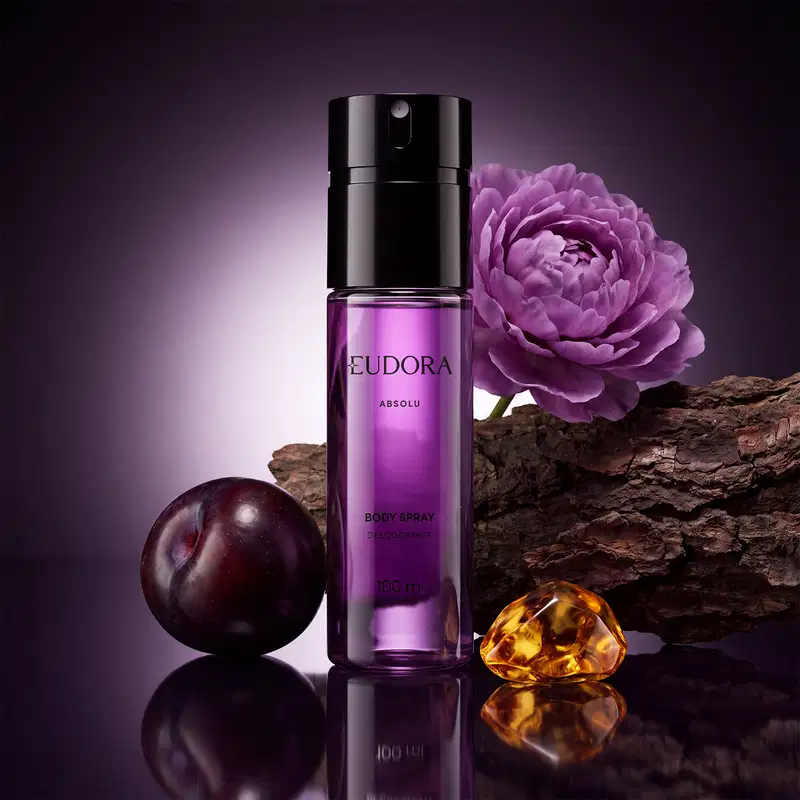 Frasco transparente, com líquido roxo claro e tampa preta, do item de perfumaria Eudora Absolu. Com uma flor de peônia roxa e ameixa preta ao redor, além de casca de Sândalo e pedra de âmbar dourado, indicando os ingredientes olfativos.