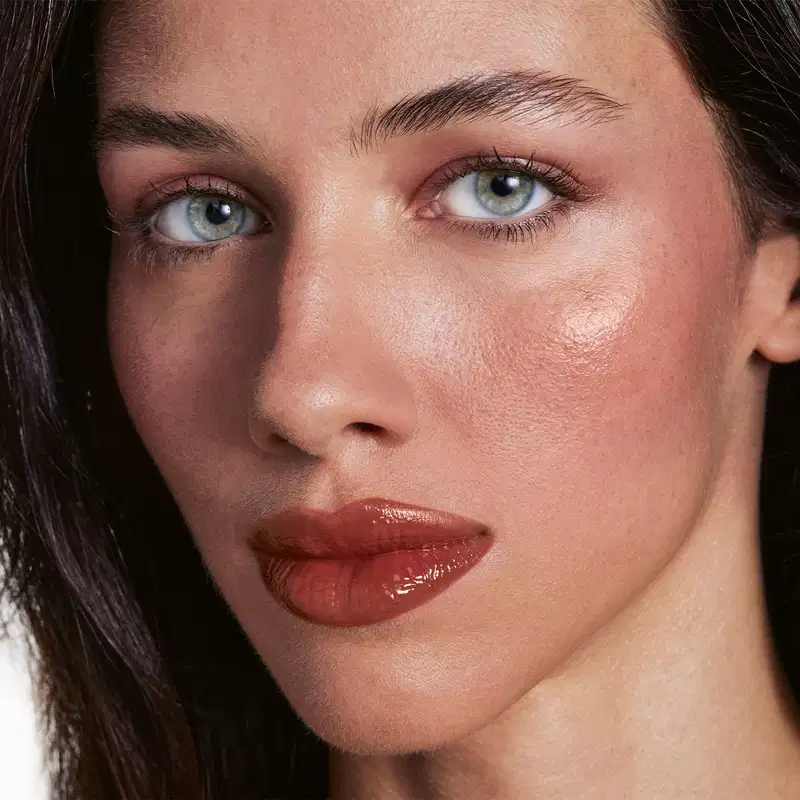 Retrato de uma mulher com olhos azuis e cabelos escuros, em close-up. A iluminação suave e o foco nos olhos e lábios destacam a beleza natural da modelo, sugerindo um ambiente de maquiagem ou cuidados de beleza.