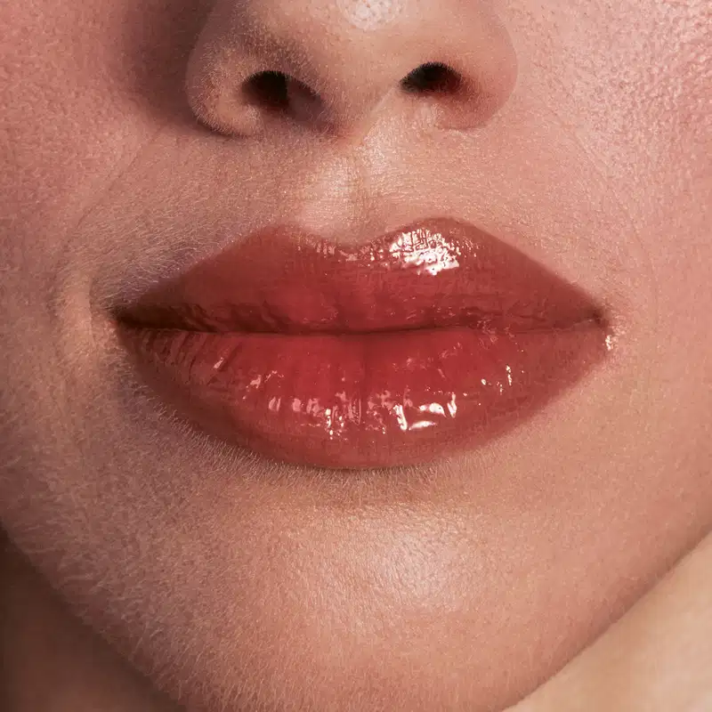 Imagem de close-up de lábios femininos com brilho labial vermelho, em um ambiente de beleza e cuidados com a pele. A textura da pele e a iluminação suave criam um cenário de cuidado e tratamento estético.