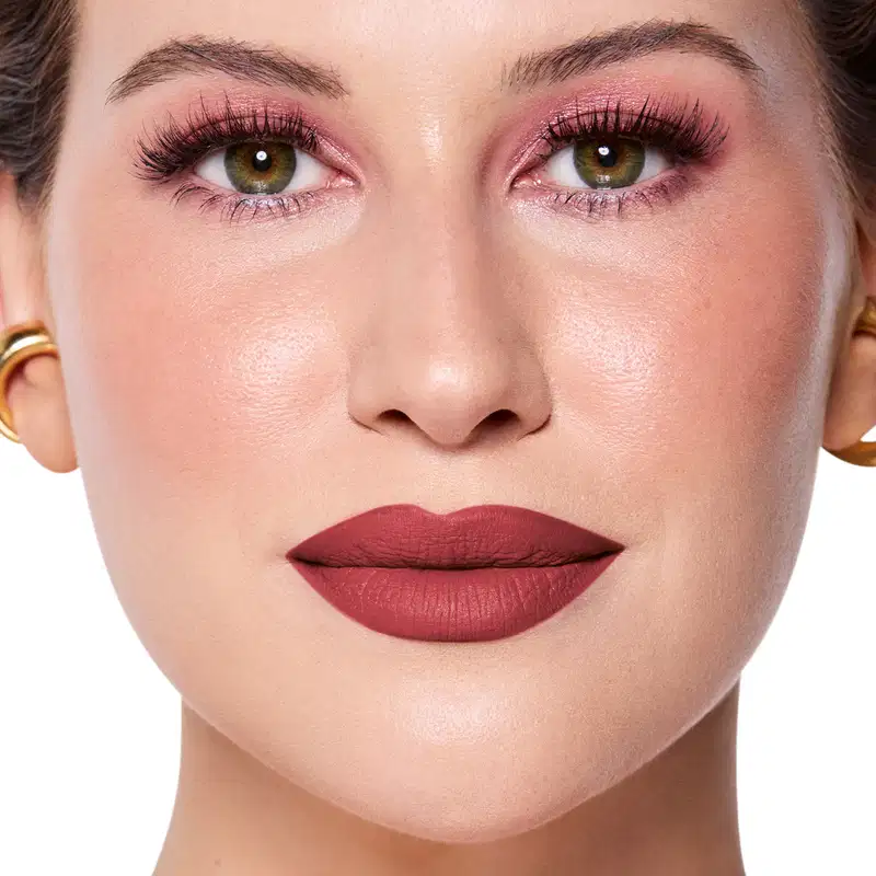 Rosto da Niina em foco, com maquiagem rosada e batom vermelho semi-matte. Olhos verdes destacados por cílios longos e sobrancelhas bem definidas. Fundo claro e iluminado, sugerindo um ambiente de beleza ou cosmética.
