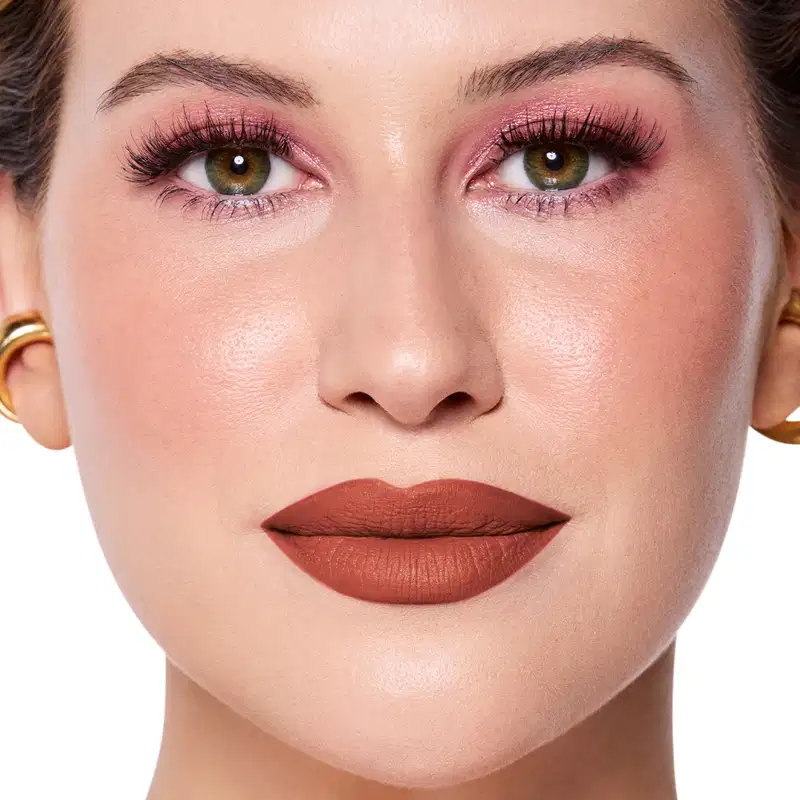 Rosto da Niina em foco, com maquiagem rosada e batom castanho semi-matte. Olhos verdes destacados por cílios longos e sobrancelhas bem definidas. Fundo claro e iluminado, sugerindo um ambiente de beleza ou cosmética.
