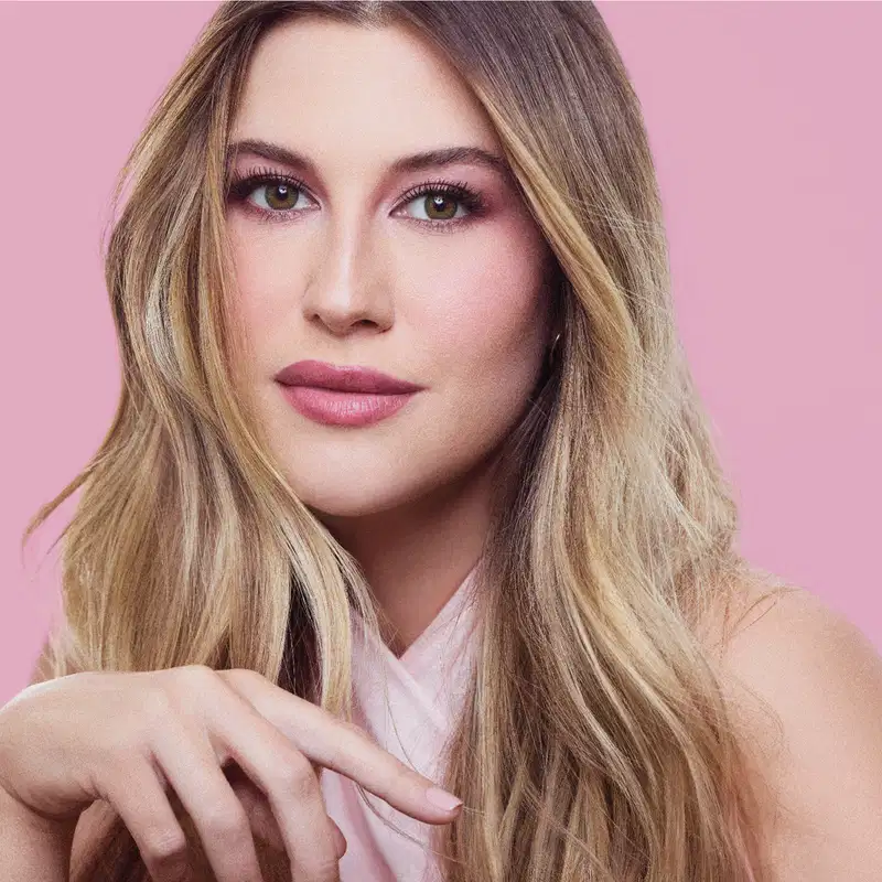 Mulher com cabelo louro comprido, maquiagem com sombra rosa e batom rosa, sobre fundo rosa uniforme, com ombro e braço visíveis.