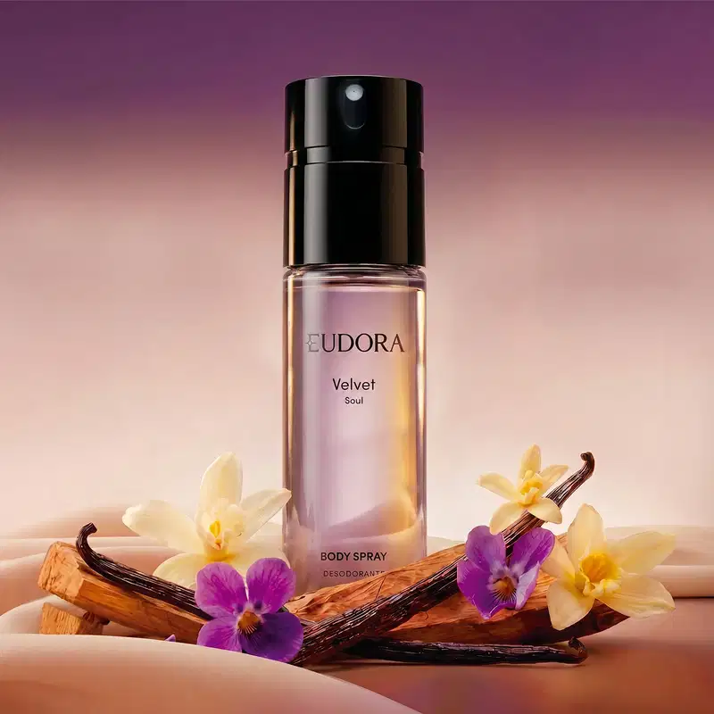 Recipiente de item de perfumaria da marca Eudora em primeiro plano, com flores de violeta e baunilha em um cenário com tons quentes de laranja e roxo. O ambiente sugere um contexto de relaxamento e bem-estar.