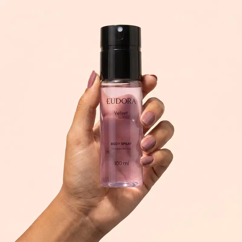 Mão segurando um frasco preto e roxo de item de perfumaria da marca Eudora, com rótulo "Velvet Soul" e informações sobre o produto. Fundo liso em tom de pêssego.