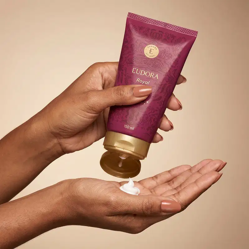 Tubo de creme da marca Eudora na cor roxa com detalhes dourados, contendo 150 ml do produto da linha Eudora Royal. O tubo é segurado por mãos femininas com unhas pintadas e podemos ver a textura do produto na mão.