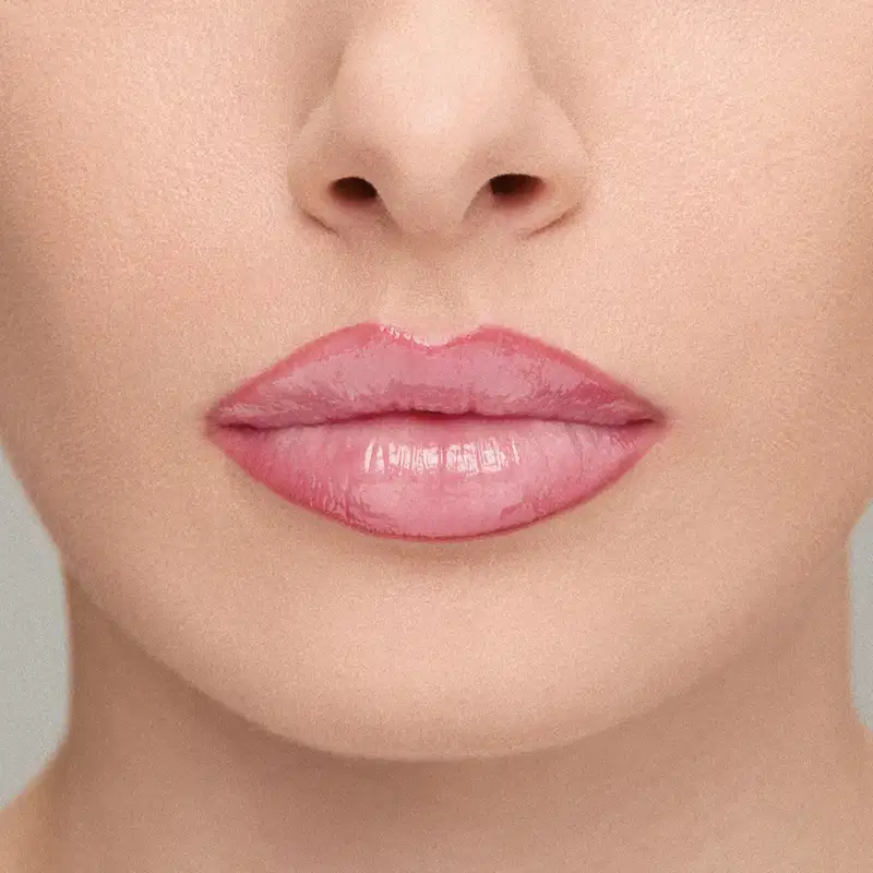 Imagem de close-up de lábios femininos com lapiseira labial e gloss rosado brilhante, em fundo claro e suave. 