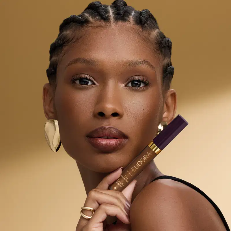 A modelo segura um corretivo líquido da marca Eudora em um ambiente de fundo dourado, destacando a maquiagem e o produto de beleza em uma imagem de alta qualidade e iluminação cuidadosa.