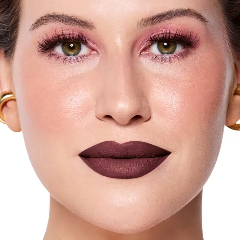 Rosto da Niina em foco, com maquiagem rosada e batom marrom escuro semi-matte. Olhos verdes destacados por cílios longos e sobrancelhas bem definidas. Fundo claro e iluminado, sugerindo um ambiente de beleza ou cosmética.
