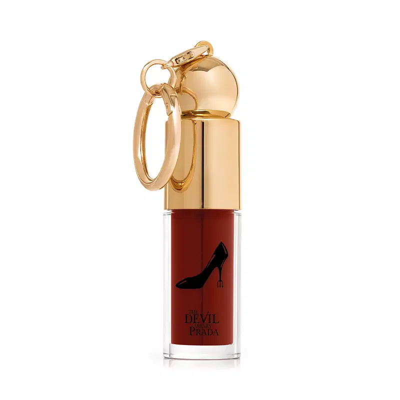 Embalagem do batom líquido, com tampa dourada e cor da make vermelho cereja. No rótulo, escrito: "The Devil Wears Prada" e imagem de um sapato de salto preto.
