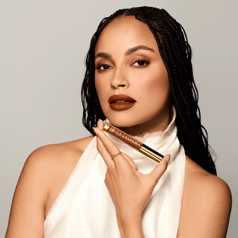 Mulher com cabelos escuros segurando o Batom Líquido Matte Supreme da marca Eudora Make em fundo branco, com iluminação suave e ambiente minimalista. Nos lábios, vemos a aplicação do batom em um tom canela.