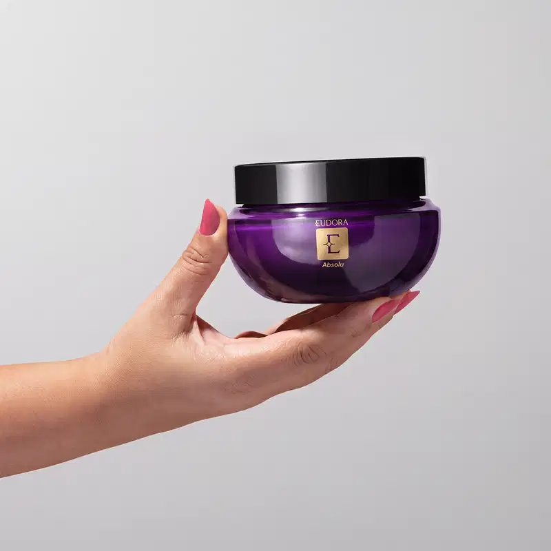 Frasco roxo com tampa preta do Creme Hidratente Desodorante Corporal da marca Eudora Absolu. O item é segurado por uma mão feminina com as unhas esmaltadas, sobre um fundo cinza.
