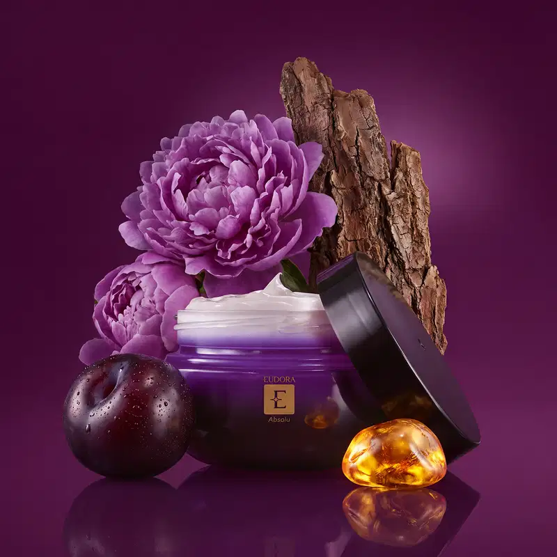 Recipiente de creme corporal roxo da marca Eudora, com flores de peônia, uma ameixa preta, pedaços de sândalo, e pedra de âmbar dourado, indicando os ingredientes que fazem parte da família olfativa. Sobre fundo roxo escuro.