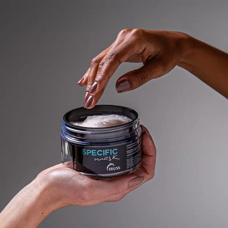 A imagem mostra uma mão segurando a máscara Truss Specific Mask. O produto tem um design elegante com o nome da marca e do produto exibidos em um rótulo escuro.